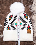 The Aztec Pom Beanie, shown on a cowhide backdrop with a ruler indicating it measures about 10 inches tall.