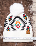 The Aztec Pom Beanie, shown on a cowhide backdrop with a ruler indicating it measures about 9 inches across.