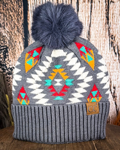 The Aztec Pom Beanie displayed on a wooden table with a wood background.
