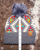 The Aztec Pom Beanie, shown on a cowhide backdrop with a ruler indicating it measures about 10 inches tall.