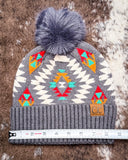 The Aztec Pom Beanie, shown on a cowhide backdrop with a ruler indicating it measures about 9 inches across.