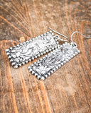 Silver rectangular earrings with intricate designs on a wooden surface