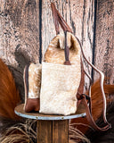 Side view of the Ranchera Hair on Hide Backpack displayed on a wooden table with wheat behind it, sitting in front of a wood back drop.