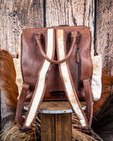 Back view of the Ranchera Hair on Hide Backpack displayed on a wooden table with wheat behind it, sitting in front of a wood back drop.