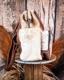 Side view of the Ranchera Hair on Hide Backpack displayed on a wooden table with wheat behind it, sitting in front of a wood back drop. 