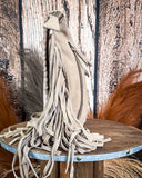 Beige leather bag with long fringes on a wooden stand against a textured wall.