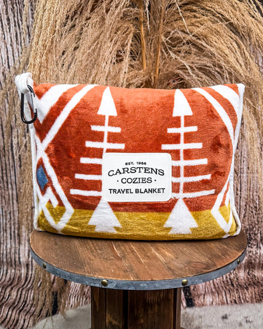 Travel blanket with geometric pattern on a wooden stool against a textured wall.