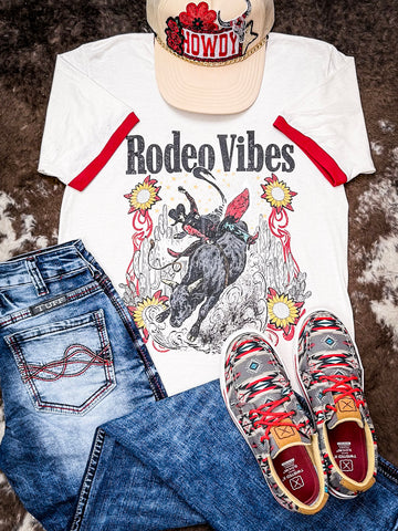 Flat lay Rodeo Vibes graphic tee with jeans, Howdy trucker hat, and Aztec sneakers