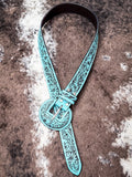 Tooled turquoise leather western belt with large floral buckle, styled on cowhide rug.