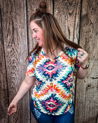 A woman wearing a colorful short-sleeve V-neck top with a vibrant geometric mandala pattern in pink, orange, blue, and yellow on a cream background, paired with blue jeans, standing against a rustic wooden plank wall.