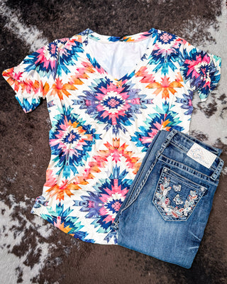 A short-sleeve V-neck top with a colorful geometric starburst pattern in pink, blue, orange, and purple on a white base, styled flat-lay with folded blue jeans on a brown and white faux fur surface.