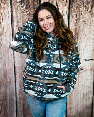 A woman wearing a Southwestern-style patterned hoodie with blue jeans, standing in front of a rustic wooden plank wall.
