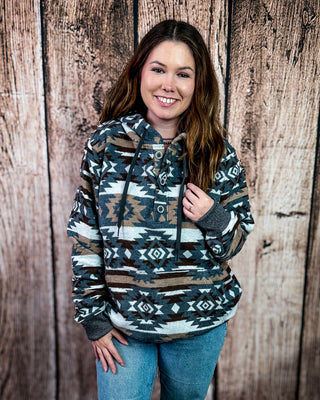 A woman smiling while wearing a long-sleeve hooded pullover with a Southwestern geometric pattern in brown, beige, gray, and white, paired with blue jeans, standing against a rustic wooden plank wall.