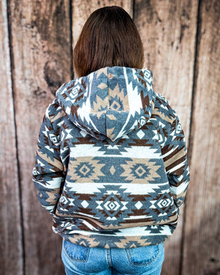 A person viewed from behind wearing a hooded sweatshirt with a Southwestern geometric pattern in slate blue, brown, beige, and white, paired with blue jeans against a rustic wooden plank background.