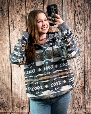 A woman takes a mirror selfie wearing a long-sleeve pullover hoodie with a Southwestern geometric pattern in gray, brown, beige, and black. She is standing in front of a rustic wooden plank wall.