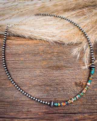 A beaded necklace with a dark metallic silver chain and a section of colorful spacer beads in orange, green, blue, and white, displayed on a rustic wooden surface with dried pampas grass.