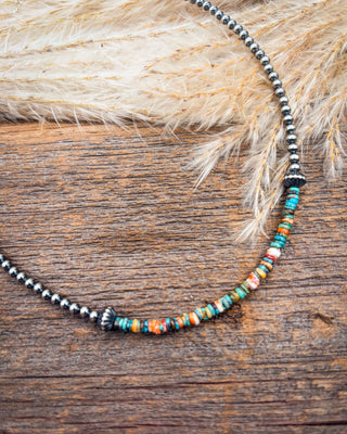 A beaded necklace with a dark metallic chain and a front section of small multicolored beads, displayed on a rustic wooden surface with dried grass.