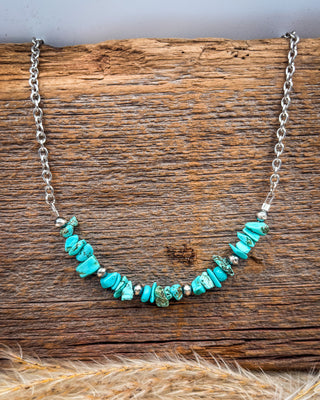 A silver chain necklace featuring a row of irregular turquoise-colored stone beads and small metallic spacer beads, displayed on a rustic wooden surface.