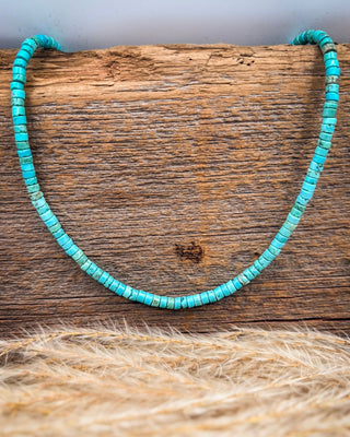A turquoise beaded necklace made of small cylindrical turquoise beads, displayed on a rustic wooden block with a blurred natural fiber surface in the foreground.