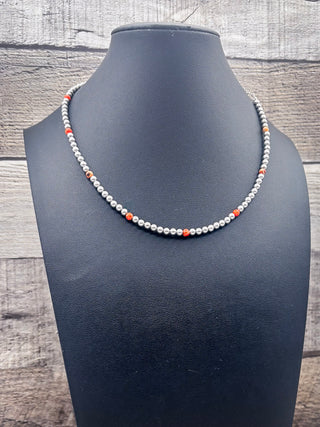 A beaded necklace displayed on a black mannequin bust, featuring small white beads with alternating orange and dark gray accent beads, set against a rustic gray wood plank background.