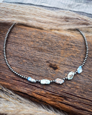 A beaded necklace with small dark metallic beads and irregular natural stone accents in light blue, white, and brown, displayed on a rustic wooden surface with tan fur.