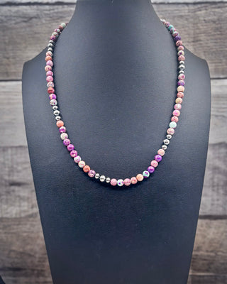 A beaded necklace displayed on a black jewelry bust against a rustic wood plank background. The necklace features round beads in pink, purple, orange, white, and metallic silver.