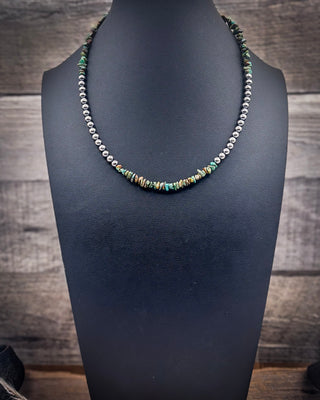 A beaded necklace displayed on a black jewelry bust against a rustic wood plank background. The necklace features a dual-design: one side is a strand of small silver beads, while the center and other side incorporate turquoise, green, and brown stone chips.
