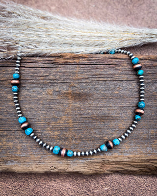 A beaded necklace with turquoise blue, dark brown, and metallic silver beads, displayed on a rustic wooden surface with dried grass.