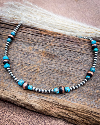 A beaded necklace with turquoise blue beads, dark brown beads, and metallic silver and bronze beads, displayed on a rustic piece of wood with dried grass.