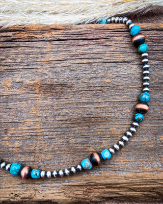 A beaded necklace with turquoise, copper, and silver beads, displayed on a rustic wooden surface with dried grass at the top.