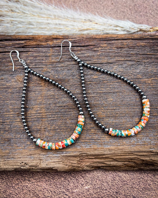 A pair of teardrop-shaped dangle earrings with black seed beads and a bottom section of colorful marbled beads in orange, teal, and cream, displayed on a rustic wooden surface.