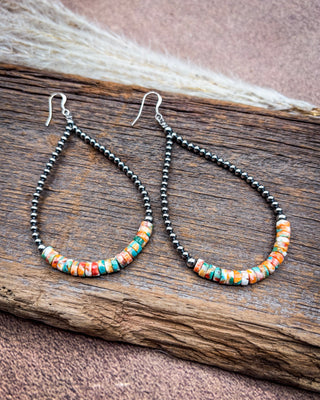 A pair of teardrop-shaped dangle earrings with black seed beads and a curved row of colorful patterned beads in orange, red, yellow, green, and blue, displayed on a rustic wooden surface.