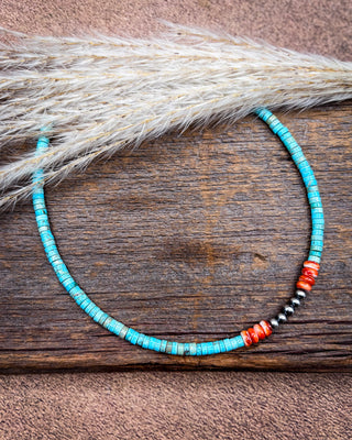 A handmade beaded necklace with turquoise-blue beads, red-orange spacer beads, and a small black-and-white bead section, displayed on a rustic wooden surface with dried grass.