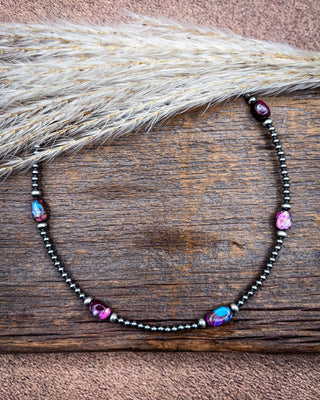 A beaded necklace with dark metallic beads and iridescent blue and pink teardrop beads, displayed on a rustic wooden surface with dried grass.