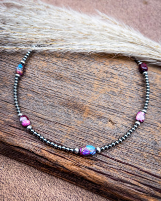 A beaded necklace with dark metallic beads, pink stones, and a central iridescent oval bead, displayed on a rustic wooden surface with dried grass.