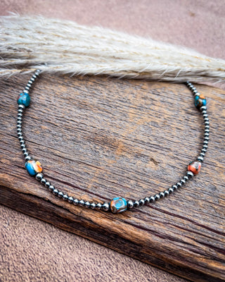 A beaded necklace with dark metallic round beads and turquoise blue, orange, and white accent beads, displayed on a rustic wooden surface with dried grass.