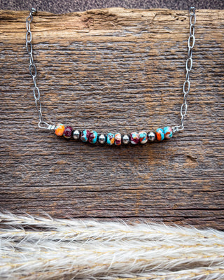 A silver chain necklace with a curved bar of colorful marbled beads, displayed on a rustic wooden surface with dried grass in the foreground.