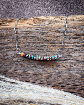A silver chain necklace with a curved bar of colorful marbled beads, displayed on a rustic wooden log with dried grass in the foreground.