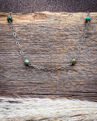 A delicate silver chain necklace with small green and brown beads, displayed on a rustic wooden surface with dried grass in the foreground.