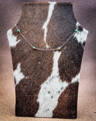 A delicate silver chain necklace with small turquoise and yellow beads, displayed on a brown and white cowhide jewelry bust against a textured brown background.
