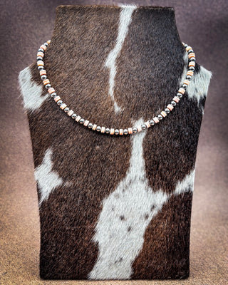 A beaded necklace with metallic rose gold, silver, and copper-toned beads displayed on a cowhide bust against a textured brown background.