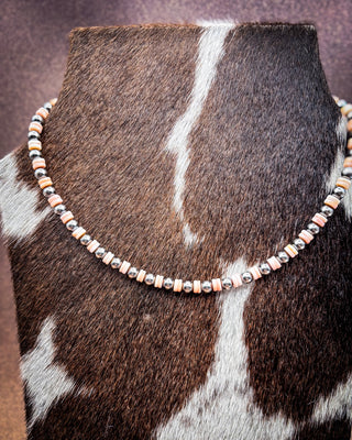 A beaded necklace displayed on a cowhide mannequin stand. The necklace features alternating metallic silver and rose gold cylindrical beads with small black accents.