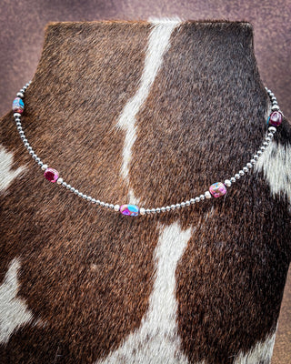 A silver beaded necklace with pink and iridescent blue gemstone accents, displayed on a cowhide jewelry stand.