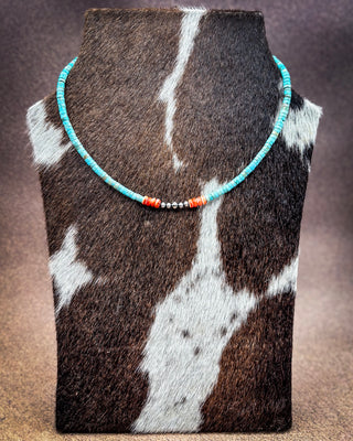 A turquoise and coral beaded necklace displayed on a cowhide jewelry bust against a warm brown gradient background.