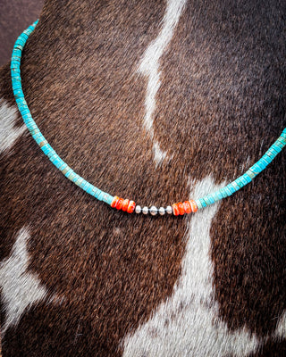 A beaded necklace draped over a cowhide surface, featuring turquoise-blue beads, coral-orange beads, and small silver spacer beads.
