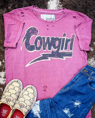 The Lightning Cowgirl Distressed Graphic Tee displayed on cowhide along with shoes and jeans.