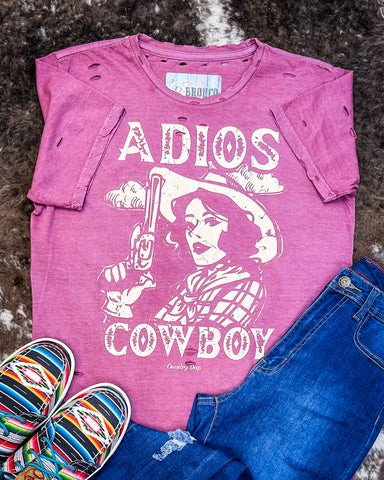 The Adios Cowboy Distressed Graphic Tee displayed on cowhide along with shoes and jeans.