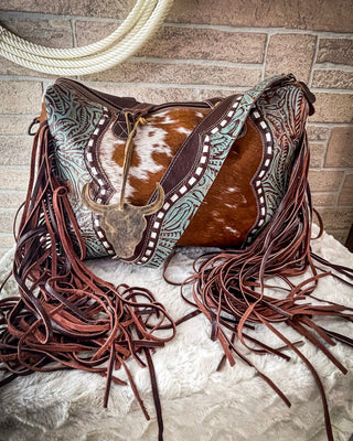 Cowhide saddle with intricate leatherwork and fringes on a stone surface.