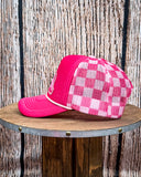 Side view of the Pink Checkered Print Barrel Racer Trucker Cap on a wood table, wood backdrop.