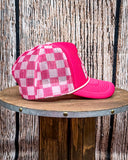 Side view of the Pink Checkered Print Barrel Racer Trucker Cap on a wood table, wood backdrop.
